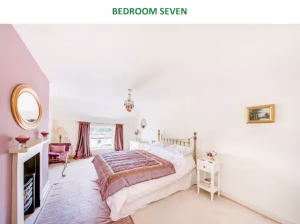 A bright and airy guest bedroom (Bedroom Seven) at Noblethorpe Hall in Silkstone, Barnsley, featuring a dusty pink accent wall, a traditional brass bed, a period fireplace, and a chaise longue by a large window.
