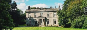 A wide panoramic daytime view of the neoclassical stone facade of Noblethorpe Hall in Silkstone, Barnsley, featuring a manicured striped lawn and lush green trees under a bright blue sky.