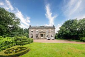 The grand neoclassical stone facade of Noblethorpe Hall in Silkstone, Barnsley, featuring a manicured lawn, circular hedge landscaping, and luxury cars parked in the private driveway.