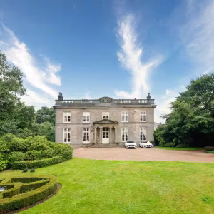 The grand neoclassical stone facade of Noblethorpe Hall in Silkstone, Barnsley, featuring a manicured lawn, circular hedge landscaping, and luxury cars parked in the private driveway.