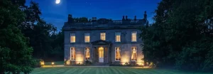 A wide panoramic night time view of the illuminated front facade of Noblethorpe Hall in Silkstone, Barnsley, under a moonlit sky with a striped green lawn.